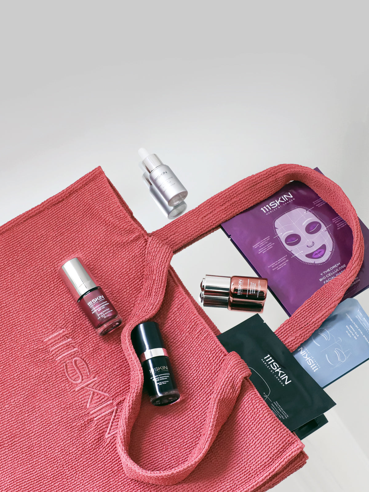 A pink 111SKIN tote bag with embossed 'ITSKIN' branding, styled with various 111SKIN skincare products including a purple sheet mask, hyaluronic acid, and other containers.