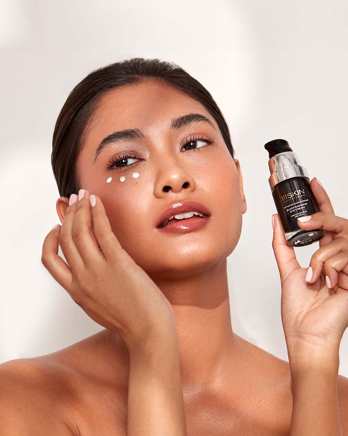 Model dabbing 3 dots of 111SKIN Eye Cream on her under eye whilst holding the glass bottle.