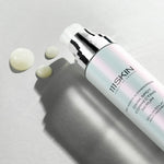 A bottle of 111SKIN Dark Spot Correcting Serum with a sleek, pearlescent white and silver design lying on a light grey surface. A few drops of the serum are dispensed nearby, showcasing its smooth, creamy texture. The lighting casts a soft shadow, adding depth to the composition.