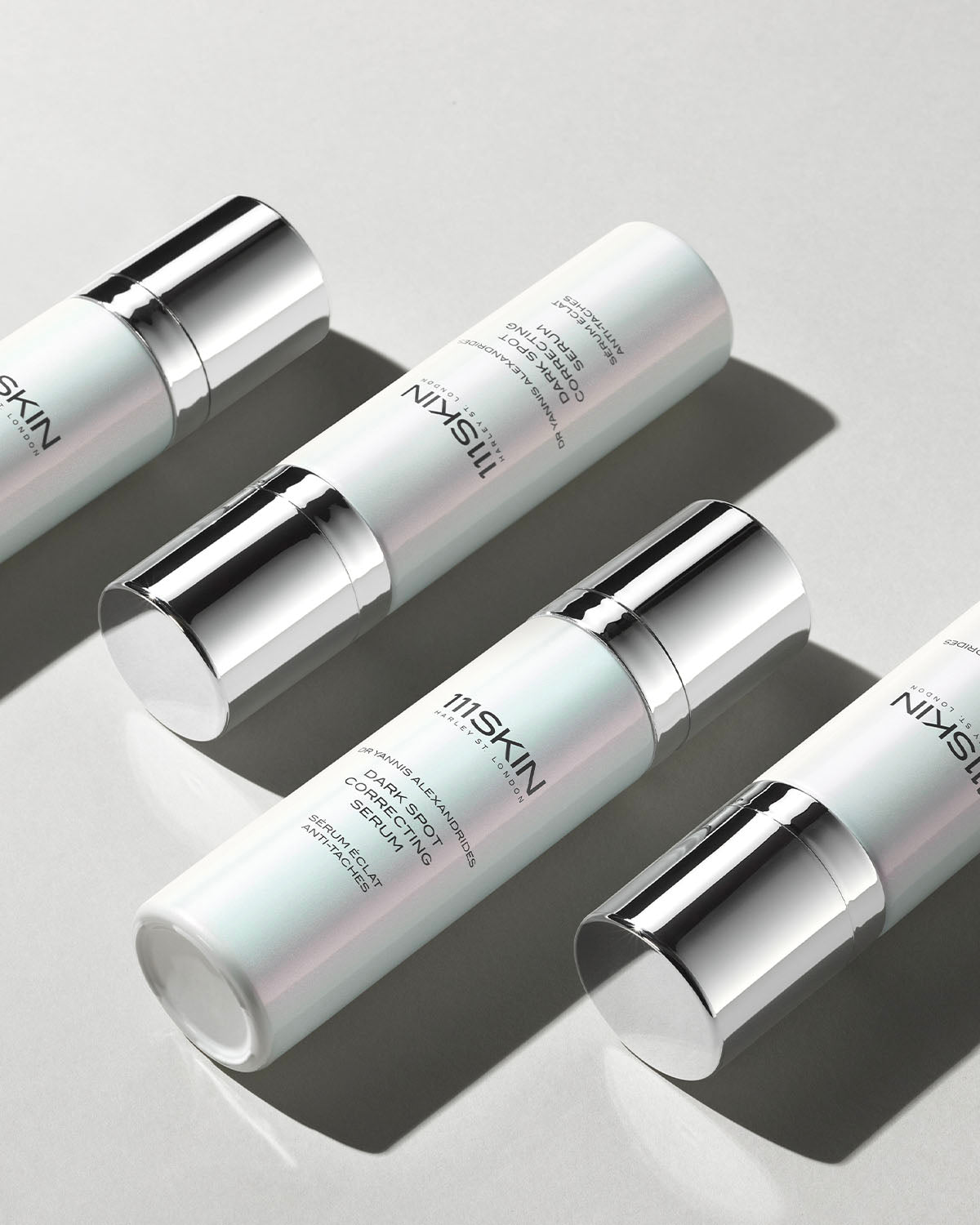 Four bottles of 111SKIN Dark Spot Correcting Serum arranged diagonally on a light grey background. The bottles have a sleek, pearlescent white design with silver caps and accents, casting soft shadows.